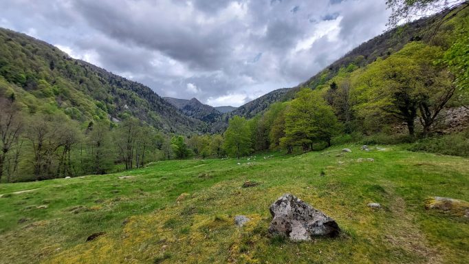 Vallée verdoyante entourée de collines, ciel nuageux et rocher au premier plan.