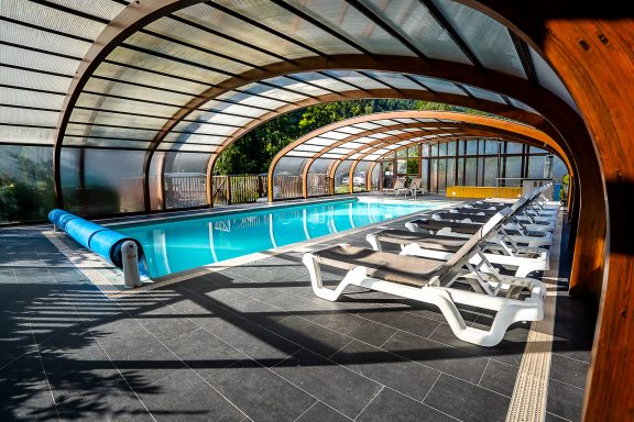 Piscine intérieure avec chaises longues et toit en verre, entourée de verdure.