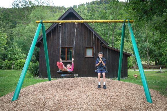 Deux enfants jouent sur une balançoire devant une maison entourée de verdure.