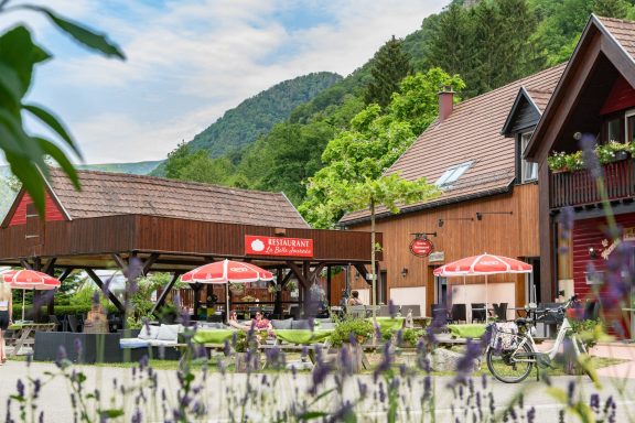 Restaurant avec terrasse, tables sous parasols, montagnes en arrière-plan et verdure.