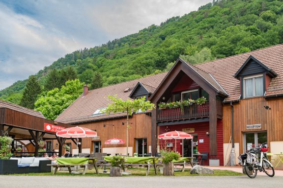 Restaurant en bois avec terrasse et parasols, entouré de verdure et de montagne.