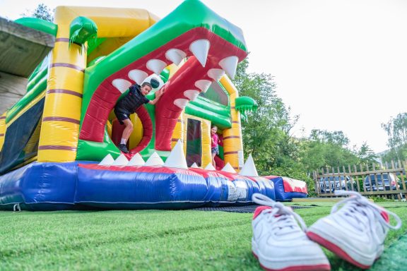 Un château gonflable en forme de dinosaure avec une personne qui y entre. Des chaussures blanches au premier plan.